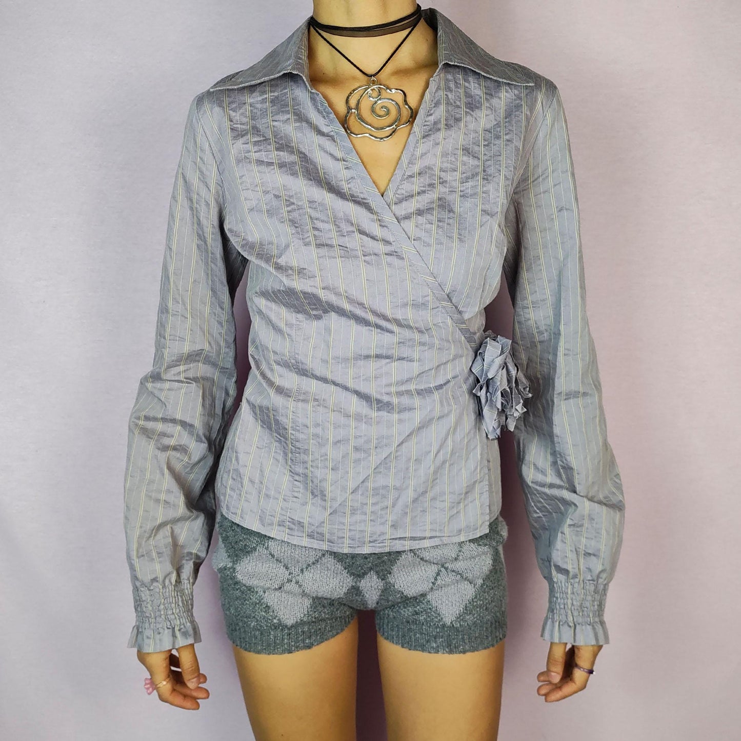 The Y2K Gray Tie Wrap Blouse is a vintage 2000s top featuring a chic wrap-around design with a side tie and delicate pinstripe pattern. Its tailored fit is complemented by a collared neckline and puffed sleeves with gathered cuffs.