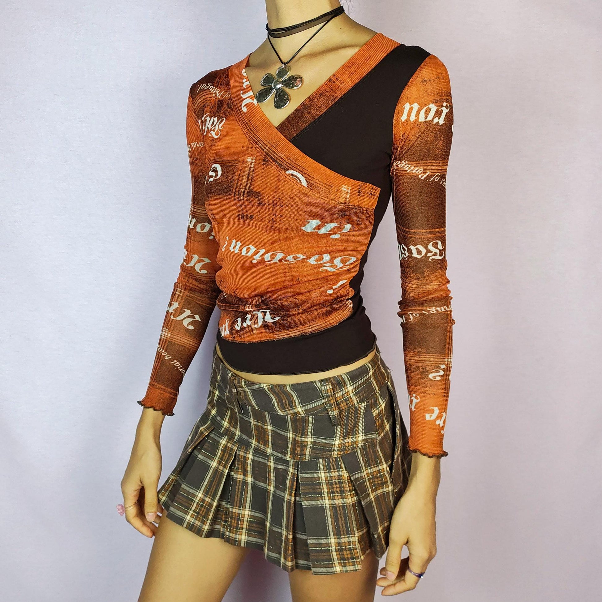 The Y2K Print Mesh Wrap Top is a vintage 2000s piece featuring a vibrant orange and brown design with bold typography prints. Crafted from a lightweight mesh fabric, it has a wrap-style front that accentuates the silhouette and long sleeves