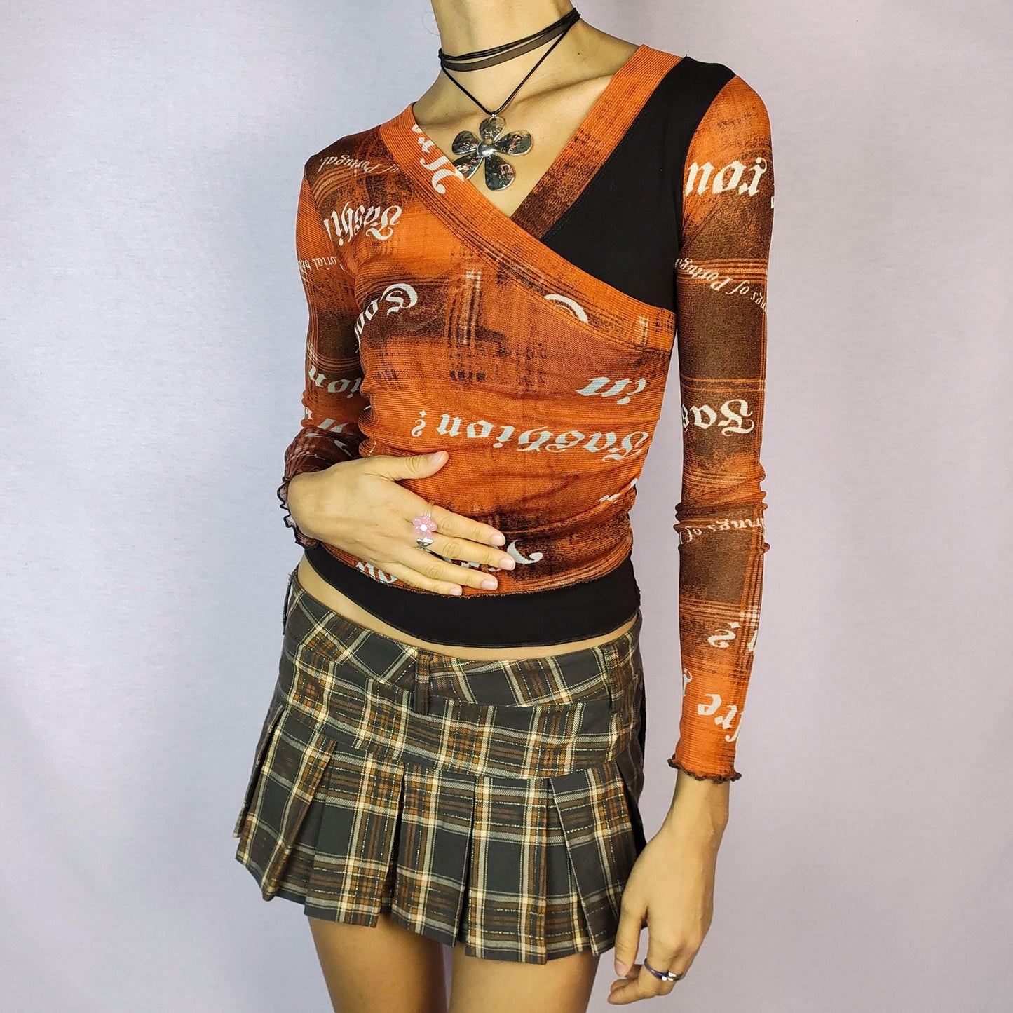 The Y2K Print Mesh Wrap Top is a vintage 2000s piece featuring a vibrant orange and brown design with bold typography prints. Crafted from a lightweight mesh fabric, it has a wrap-style front that accentuates the silhouette and long sleeves