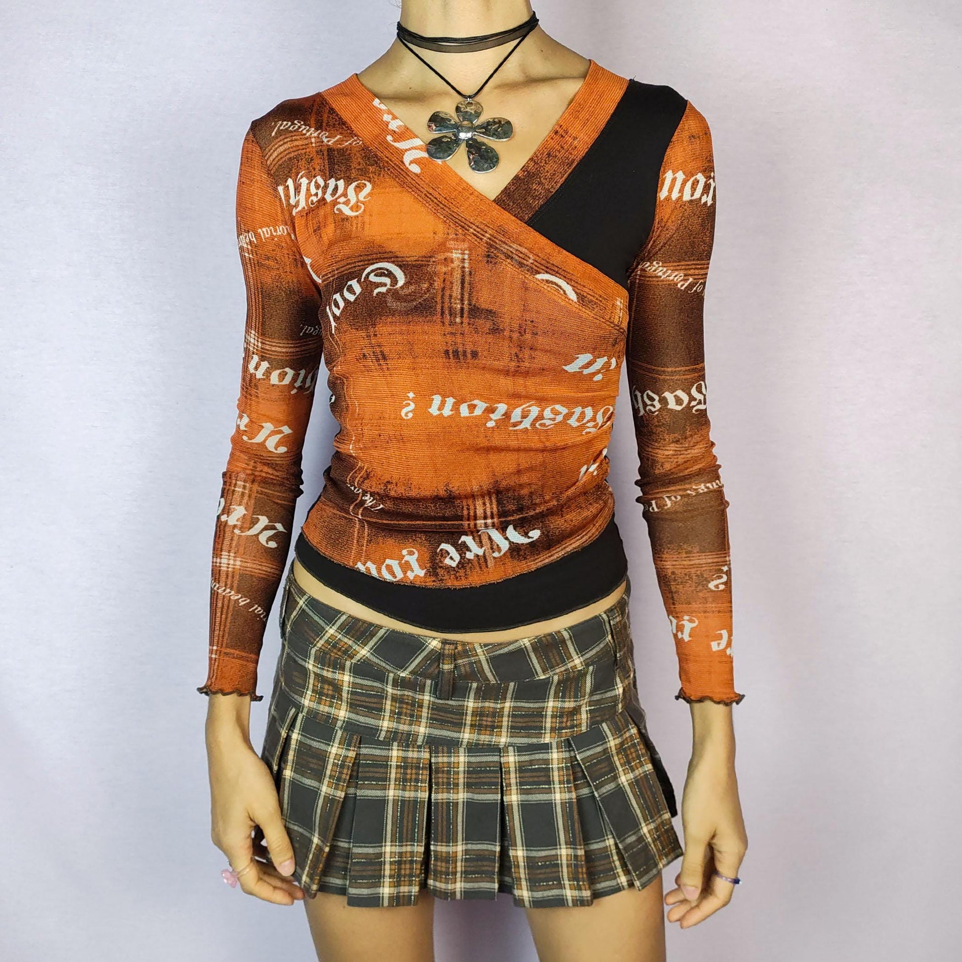 The Y2K Print Mesh Wrap Top is a vintage 2000s piece featuring a vibrant orange and brown design with bold typography prints. Crafted from a lightweight mesh fabric, it has a wrap-style front that accentuates the silhouette and long sleeves