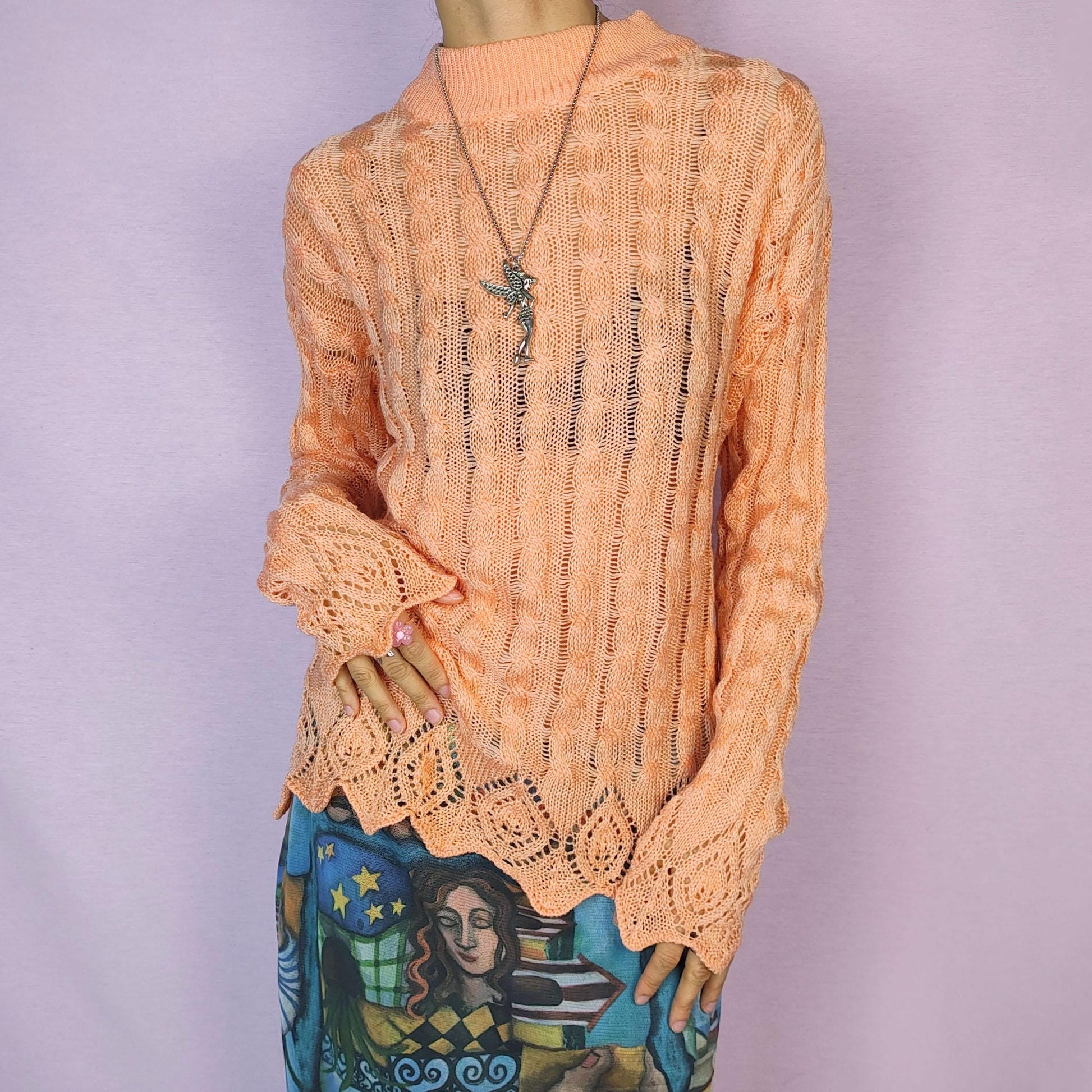 The Vintage 90s Peach Knit Sweater features a soft knit design with an openwork cable pattern, delicate scalloped hem detailing, long sleeves and a mock neckline. This lightweight sweater is crafted in a warm peach tone. Adult World Shop.