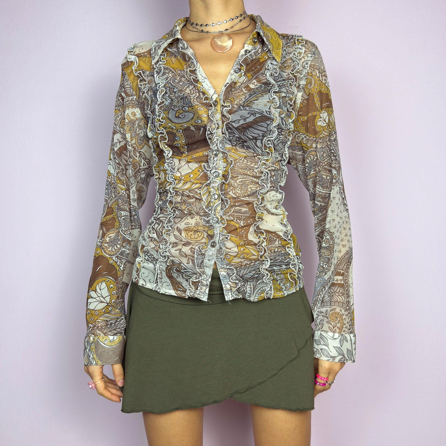 The Y2K Mesh Frill Blouse is a vintage 2000s romantic statement piece. Featuring a semi-sheer beige mesh fabric adorned with intricate paisley and floral patterns in earthy and mustard tones. adult world shop